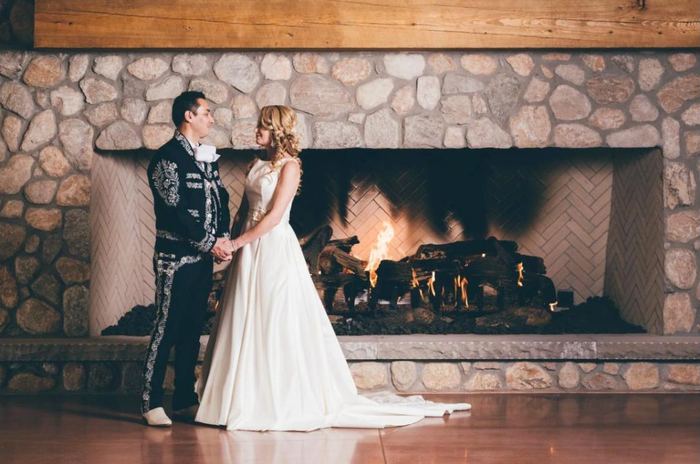 Bride and groom in charro suit holding hands by a stone fireplace